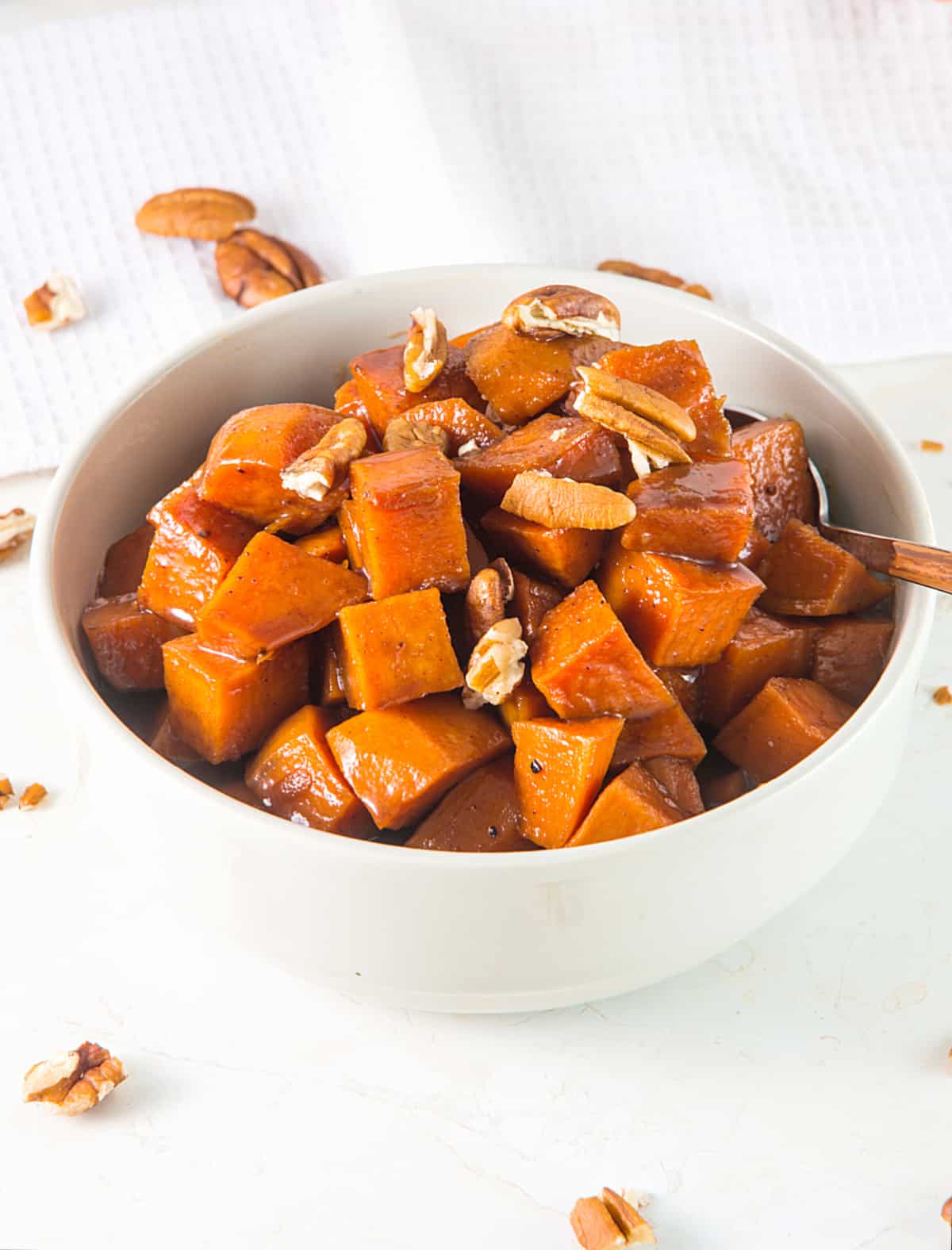 White bowl of candied sweet potatoes with pecans on a white kitchen towel.