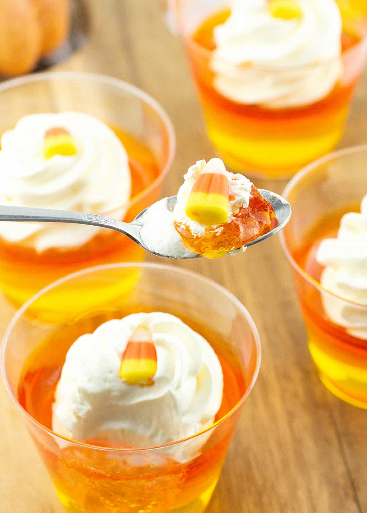 Spoon with candy corn jello shot. More jello cups beneath on a wooden surface.