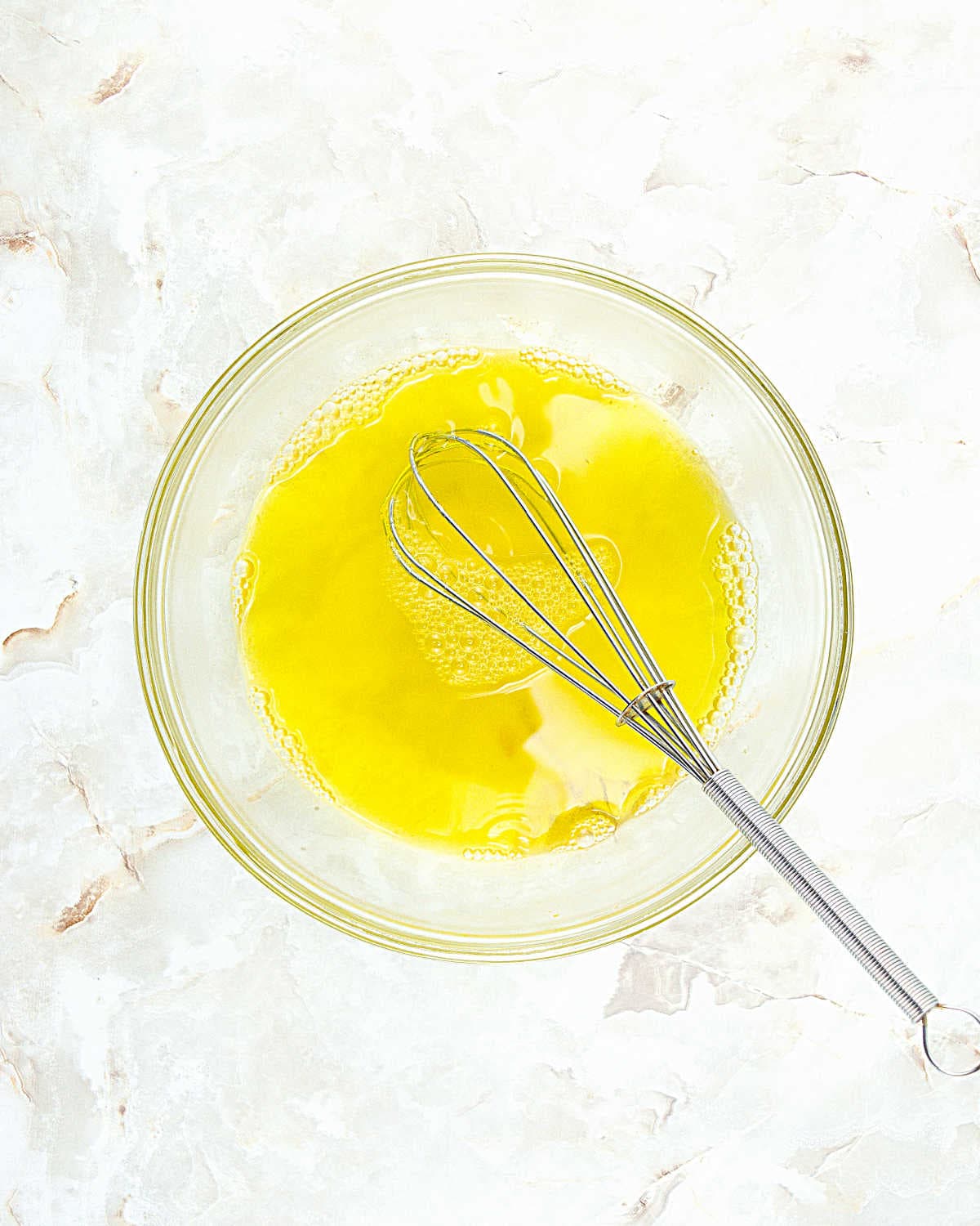 Glass bowl with yellow jello mixture and metal whisk. White beige marbled surface.