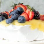 White text overlay on mixed berry lemon pavlova on a white plate. Grey background.