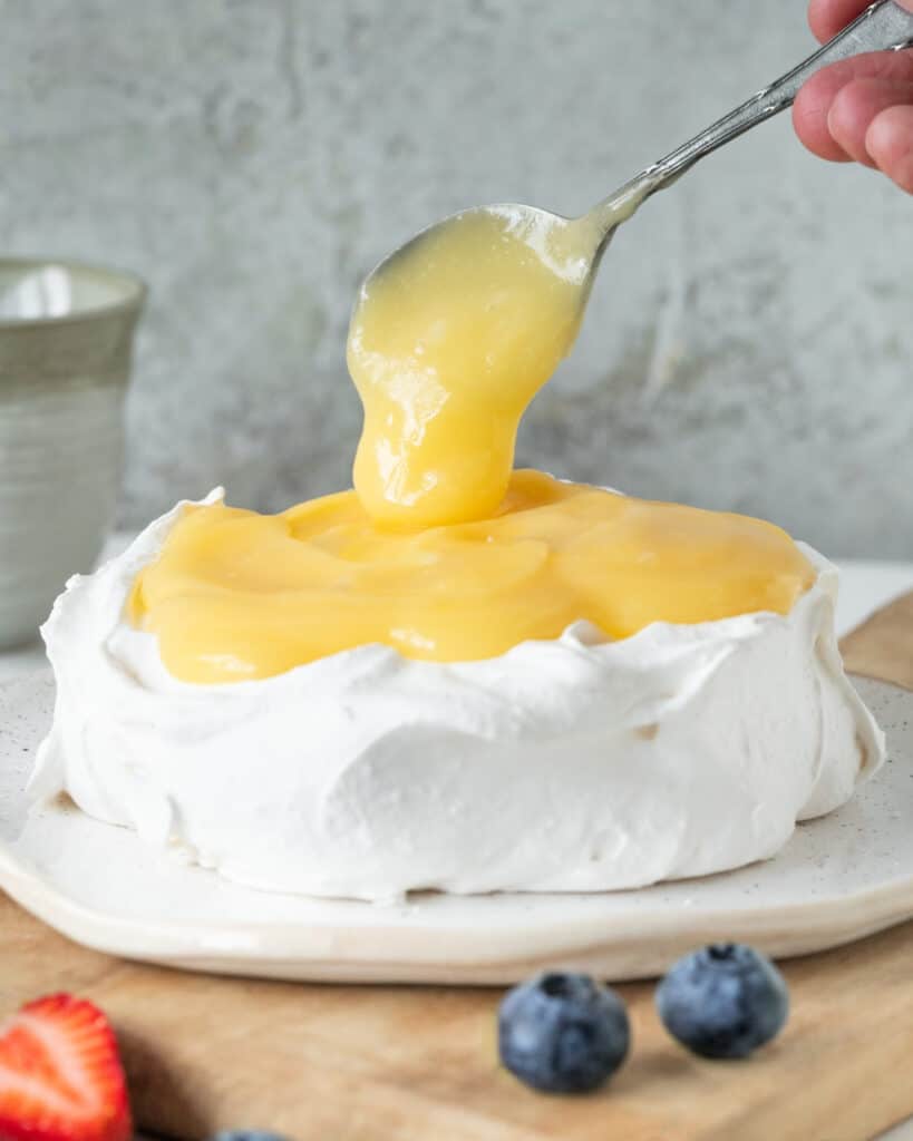 Spooning lemon curd on a merigue disc on a white plate. Grey background.