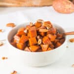 Candied sweet potatoes with pecans in a white bowl on a white surface.
