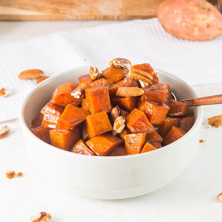 Candied sweet potatoes with pecans in a white bowl on a white surface.