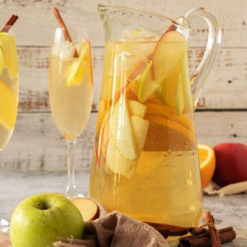 Tall pitcher and stem glasses of apple cider white wine sangria with sliced fruit. Whitish wood background.