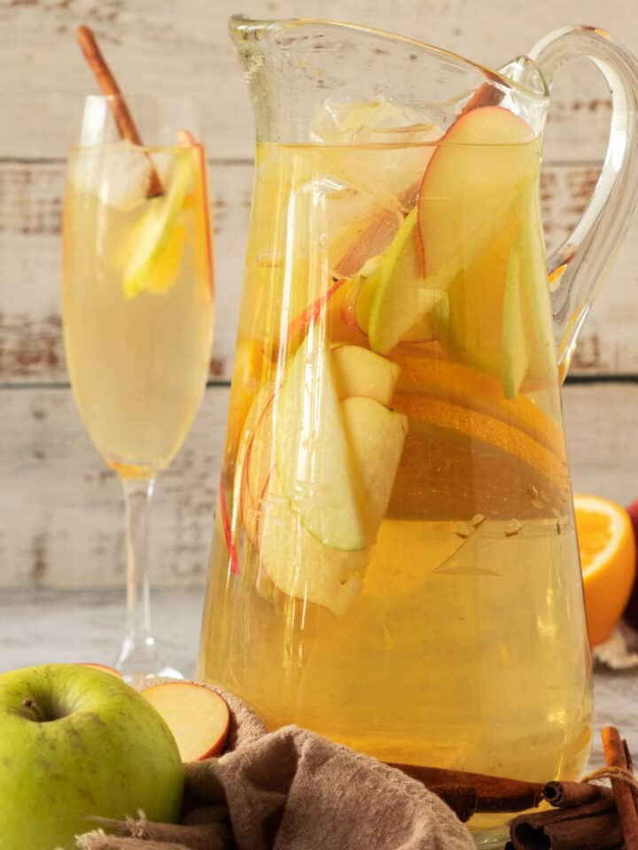 Tall pitcher and stem glasses of apple cider white wine sangria with sliced fruit. Whitish wood background.