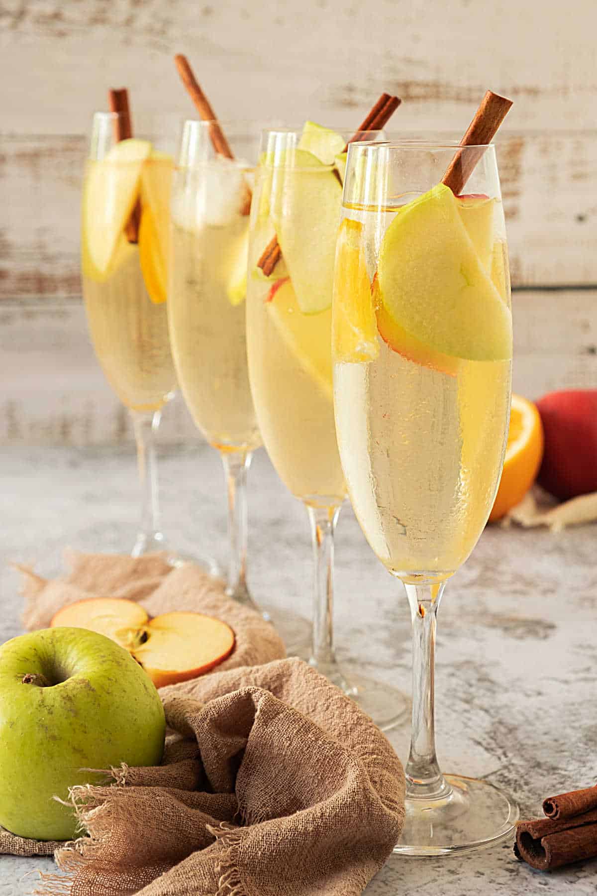 Four long stem glasses with apple sangria and cinnamon sticks. Grey background, fresh fruit, brown cloth. 