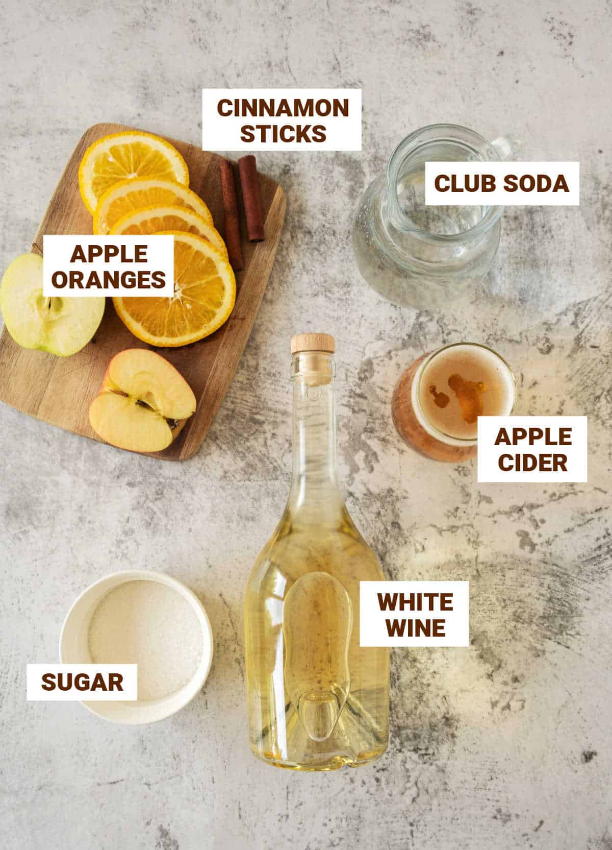 Ingredients for apple cider sangria including bottle of white wine, orange and apple slices, sugar, club soda. Grey surface. 