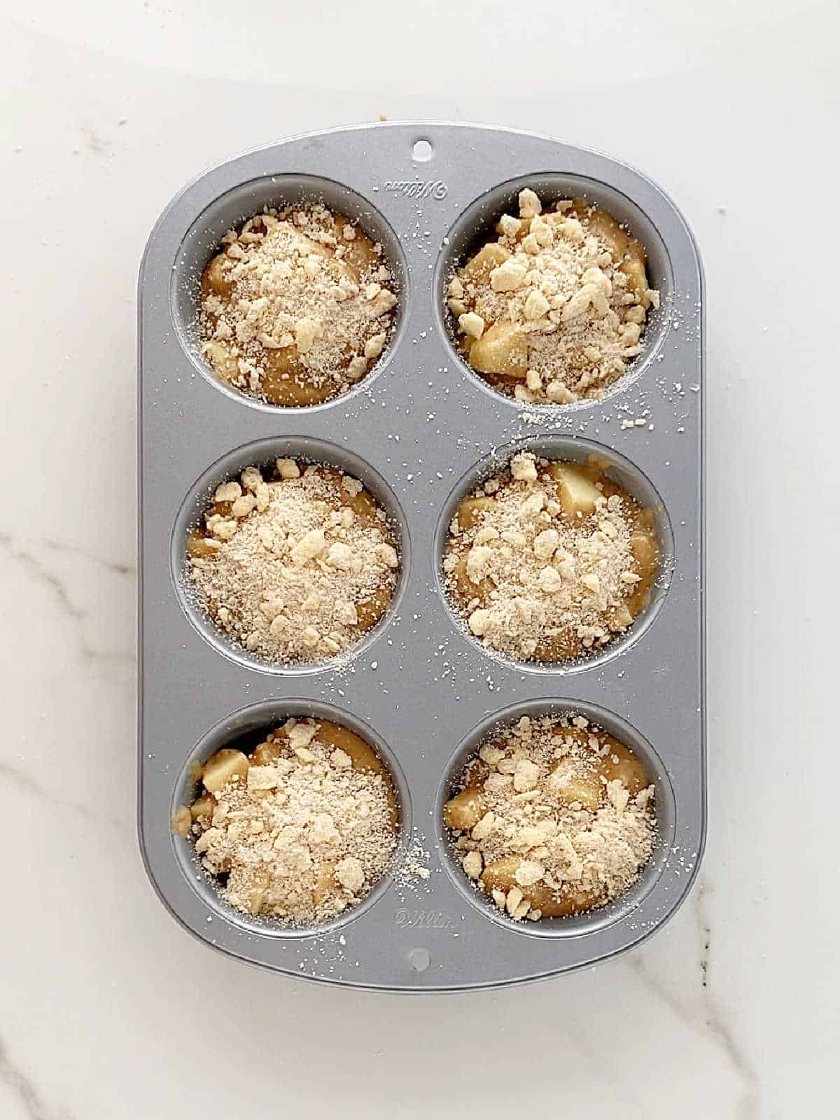 Metal jumbo muffin pan with apple crumb muffins before baking. White marbled surface.