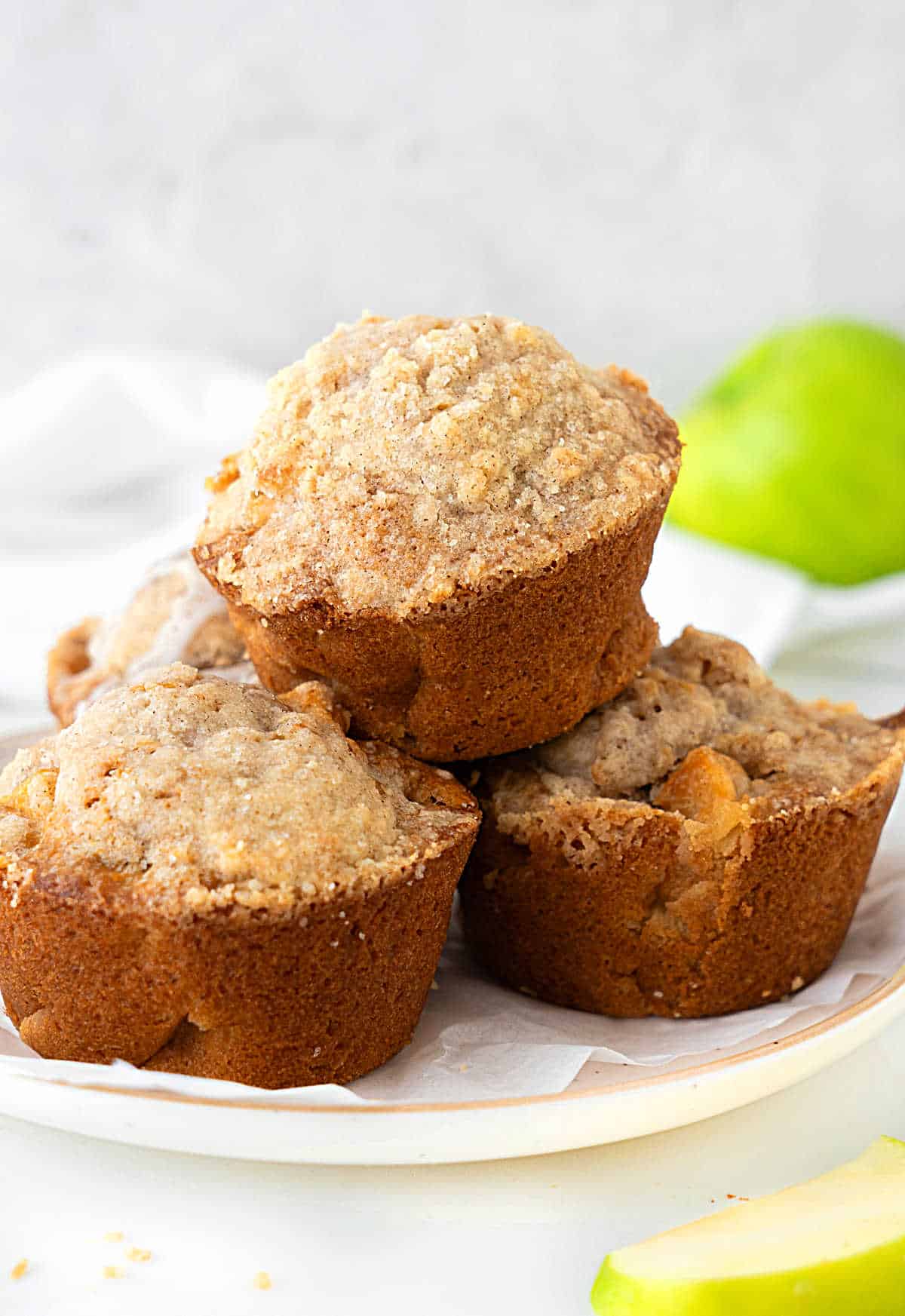 Grey and white background with green apple and white plate with pile of apple crumb muffins. 