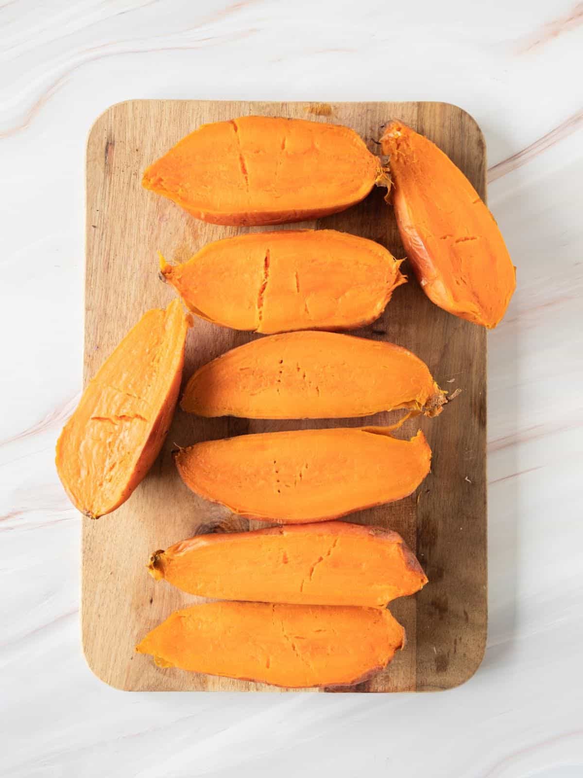 Halved sweet potatoes on a wooden board on a white marbled surface.
