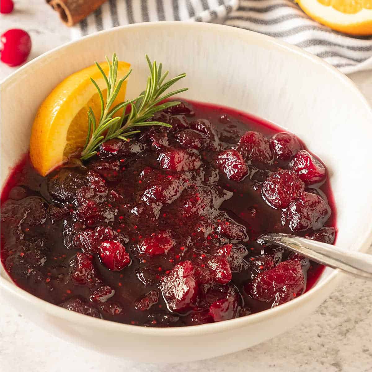 Orange cranberry sauce in a white bowl with a spoon, rosemary sprig and orange wedge.