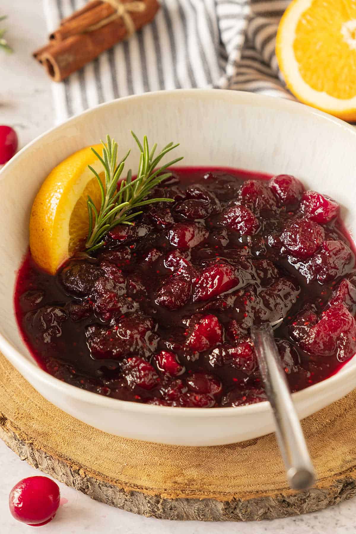 White bowl with cranberry sauce on a wooden round, orange slices, striped cloth in the background. 