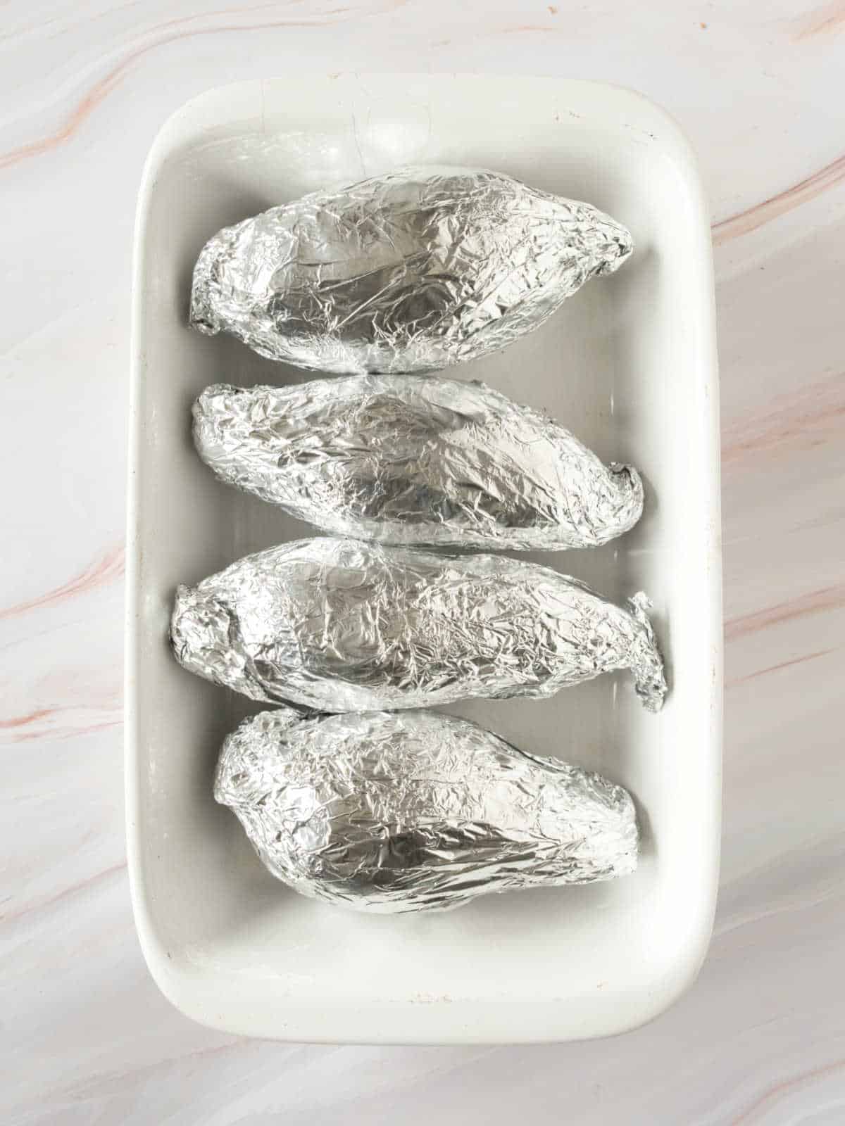 Foil wrapped sweet potatoes in a white rectangular dish on a white marbled surface.