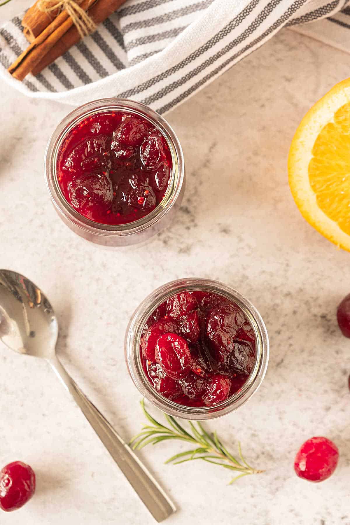 Top view of glass jars with cranberry sauce. Beige surface, striped cloth, orange pieces, silver spoon.