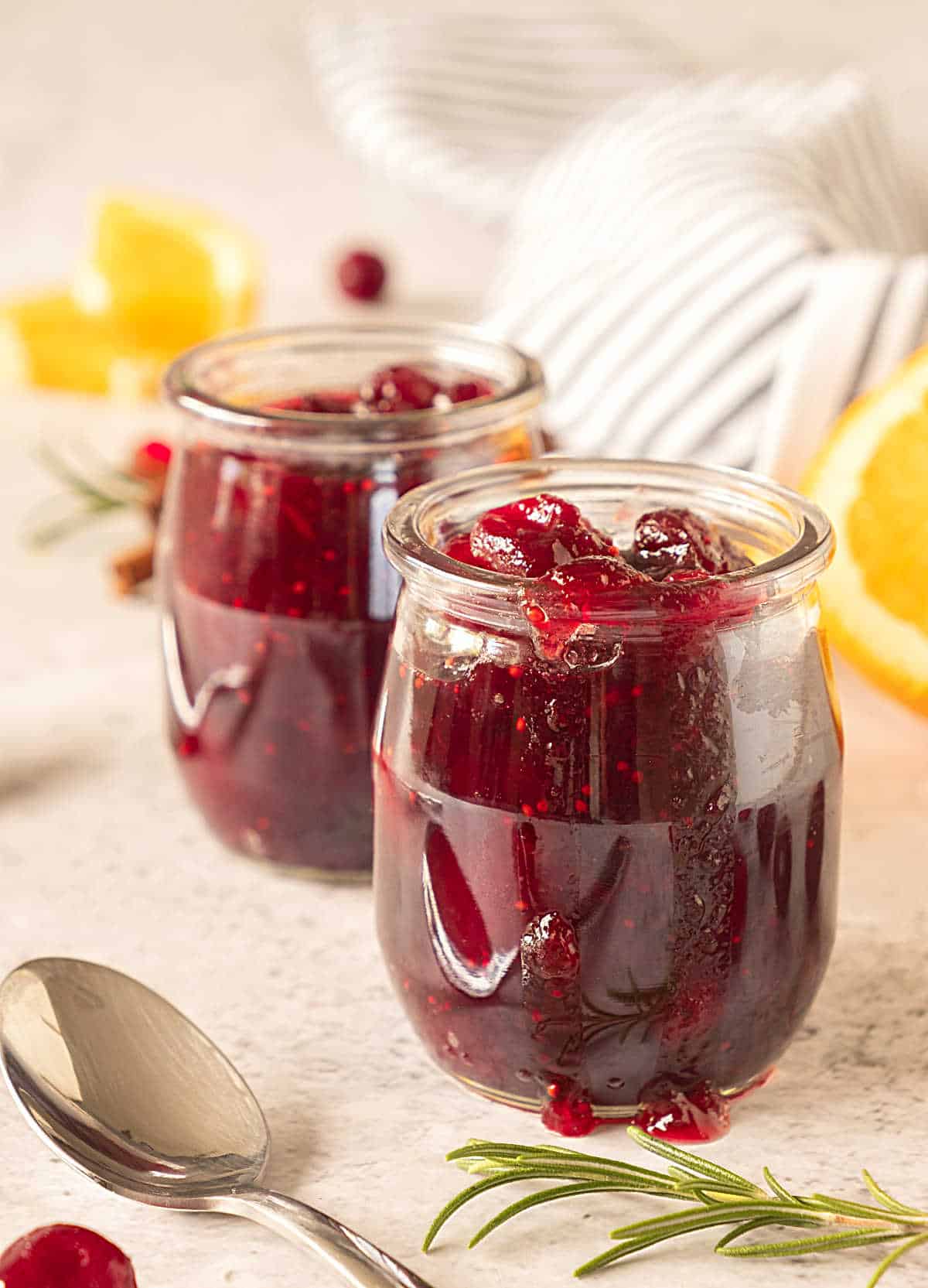 Two glass jars with cranberry sauce on a beige background with a striped cloth. Orange slices, a spoon. 