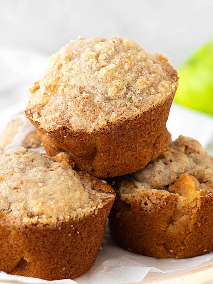 Pile of apple crumb muffins on a white plate. White background with a green apple.