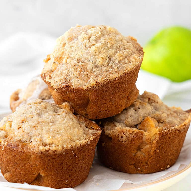 Pile of apple crumb muffins on a white plate. White background with a green apple.