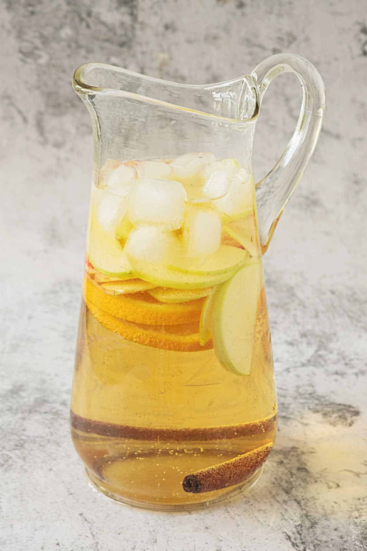 Grey background with pitcher containing apple cider sangria with ice and fruit slices.
