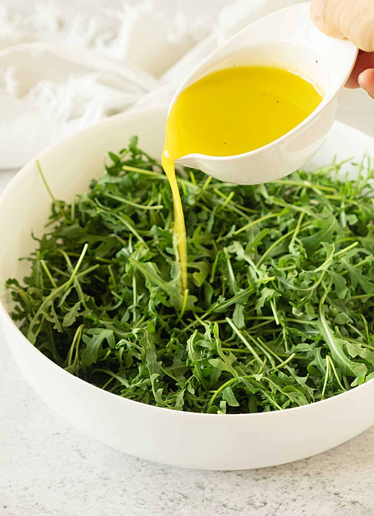 A white bowl with arugula and spinach. Dressing being poured from a white cup.