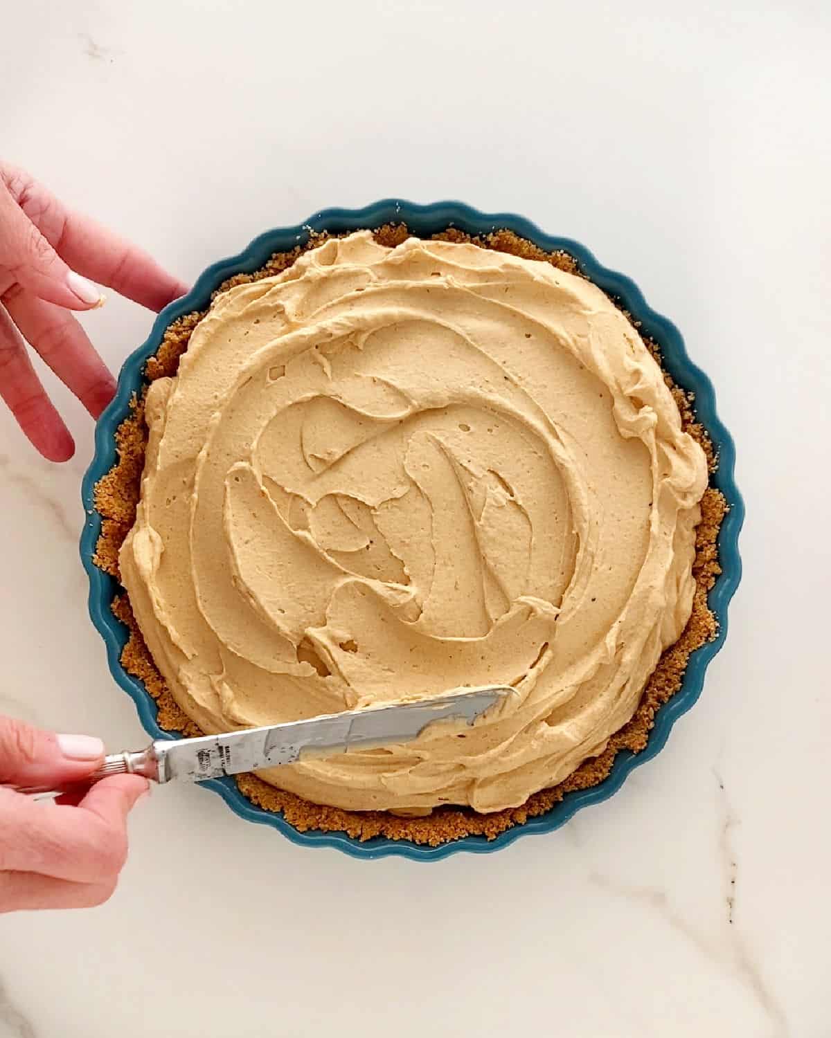 Spreading pumpkin mousse filling in a graham crust. Teal pie dish on a white surface.