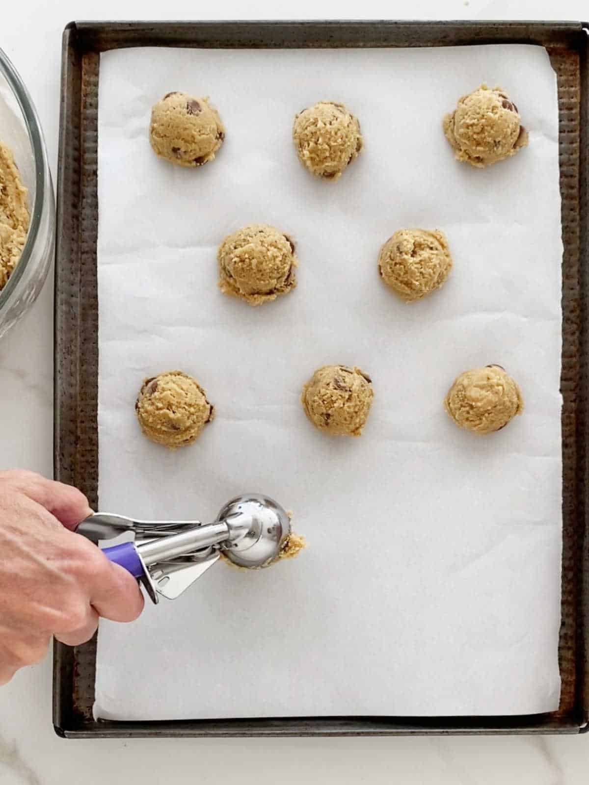 Scooping chocolate chip cookies onto white parchment paper on a dark metal sheet.