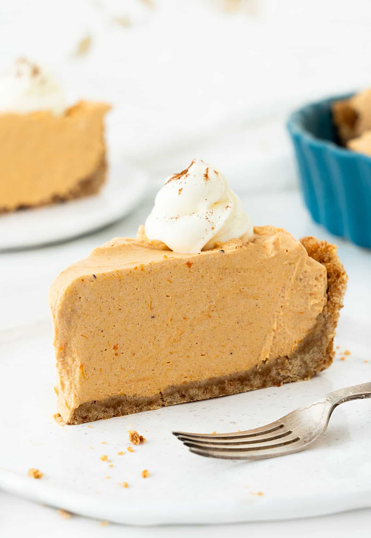 Slice of pumpkin mousse pie on a white plate. White background with more pie slices and teal dish.