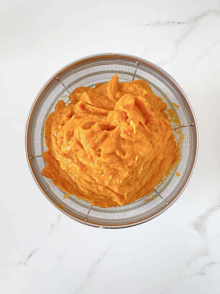Draining sweet potato puree in a metal colander. White marbled surface. Top view.