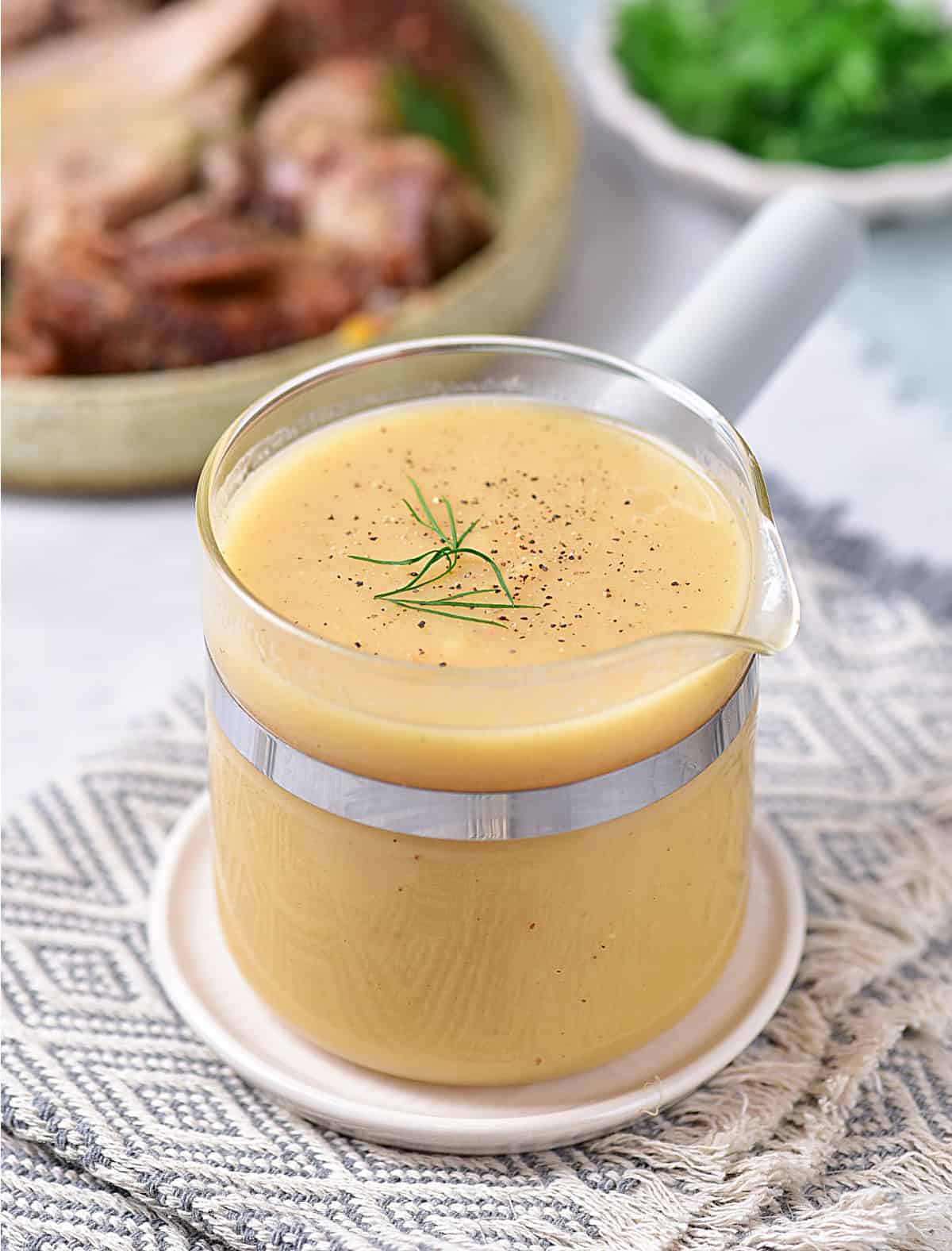 Glass pouring cup with gravy on a grey white towel. Sliced turkey and parsley in the background.