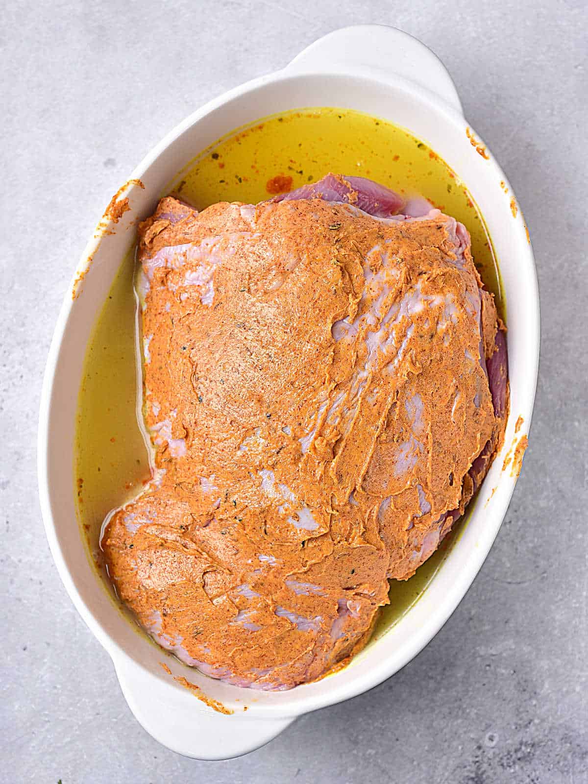 White oval dish with seasoned turkey thigh before baking. Grey surface. 