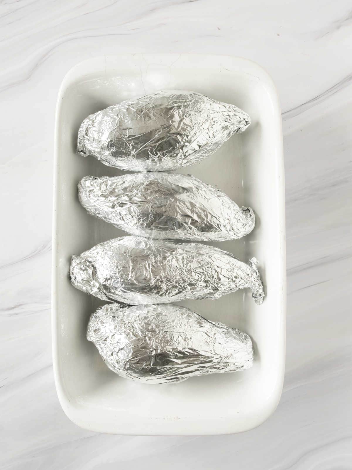 Four foil-wrapped sweet potatoes in a white baking dish on a white marbled surface.