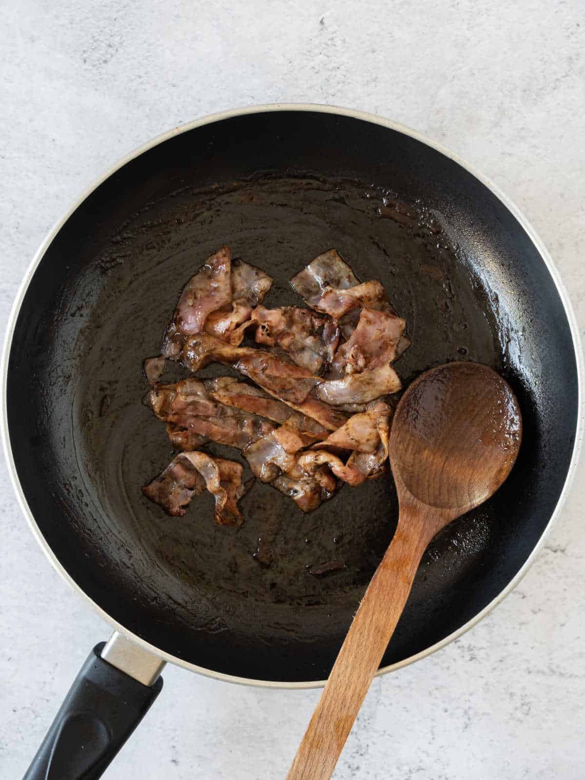 Cooked bacon strips with balsamic in a black skillet with a wooden spoon. Grey surface.