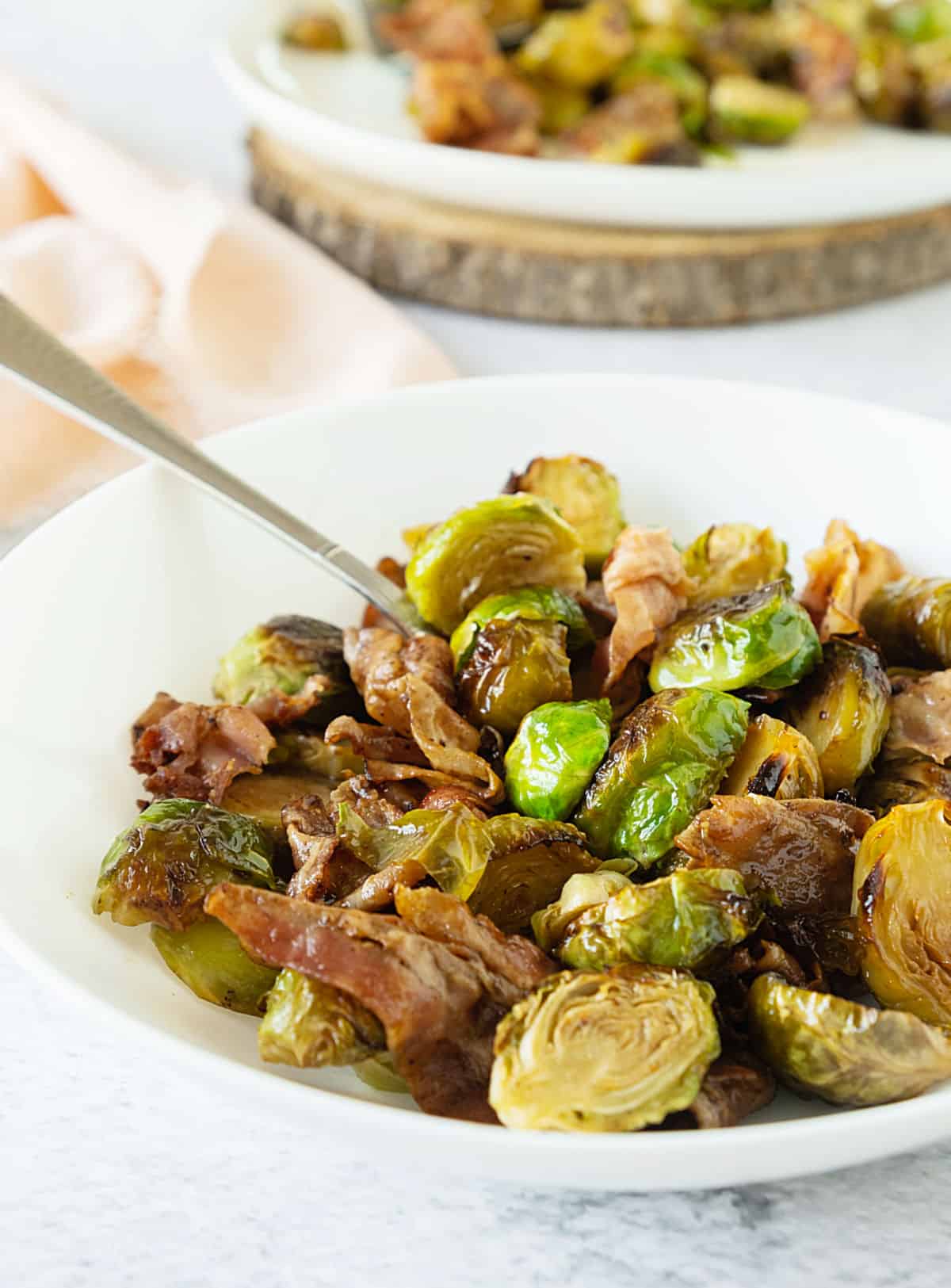 Brussels sprouts with bacon in a white bowl with silver spoon. Another plate and pink cloth in the background.