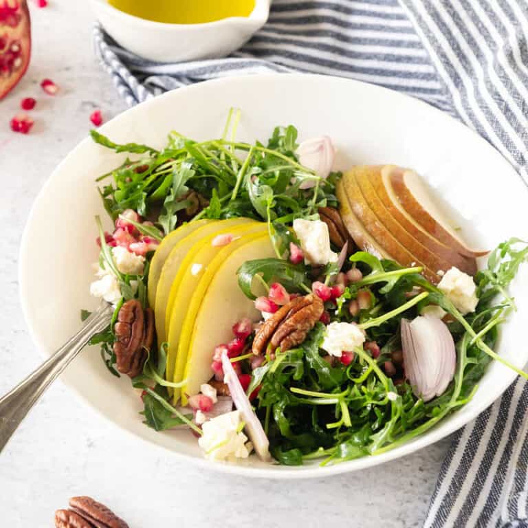 A white bowl with pear, arugula and pomegranate salad. Blue striped cloth. White surface.