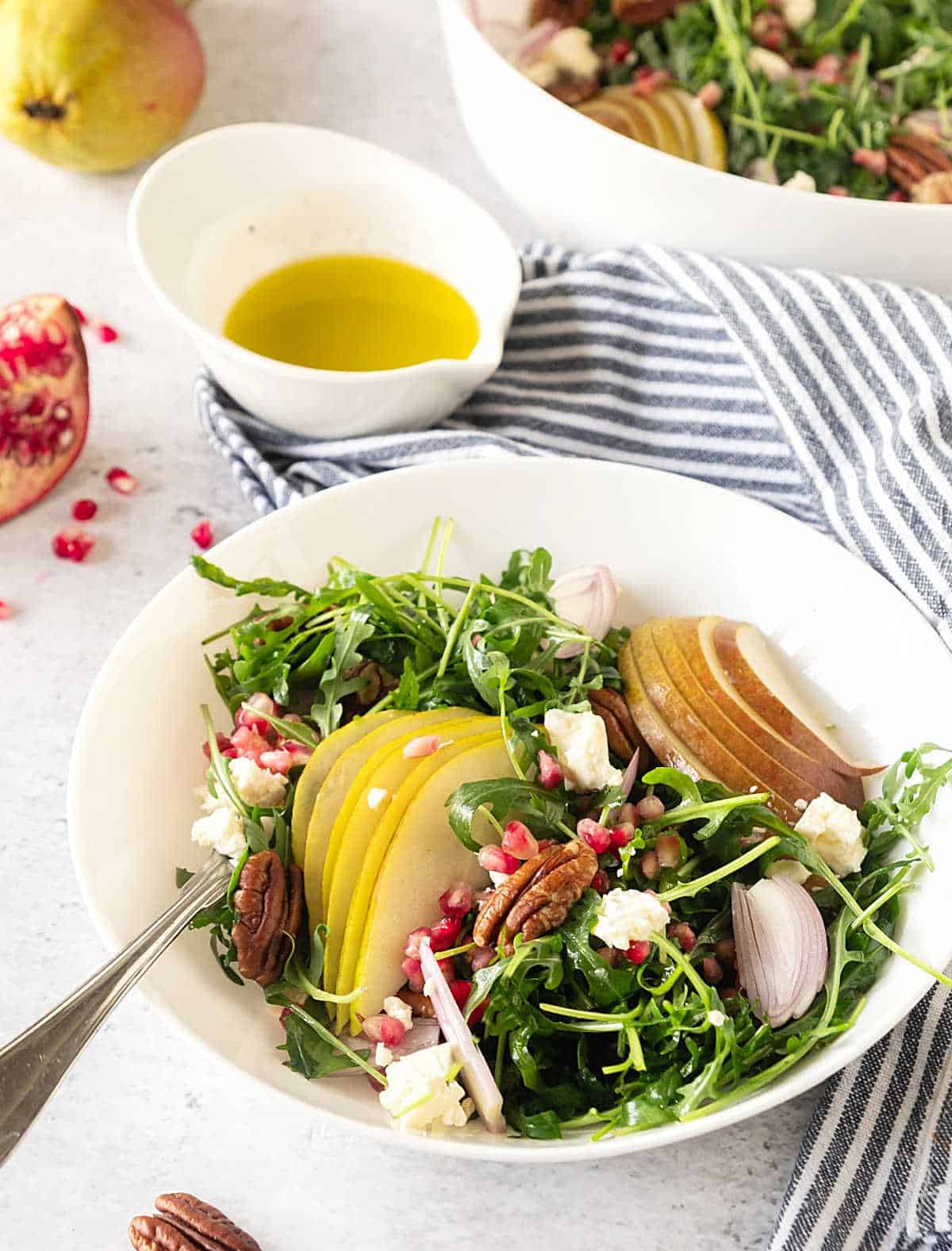 Striped blue cloth with bowls of pomegranate spinach pear salad. White bowl with dressing. 