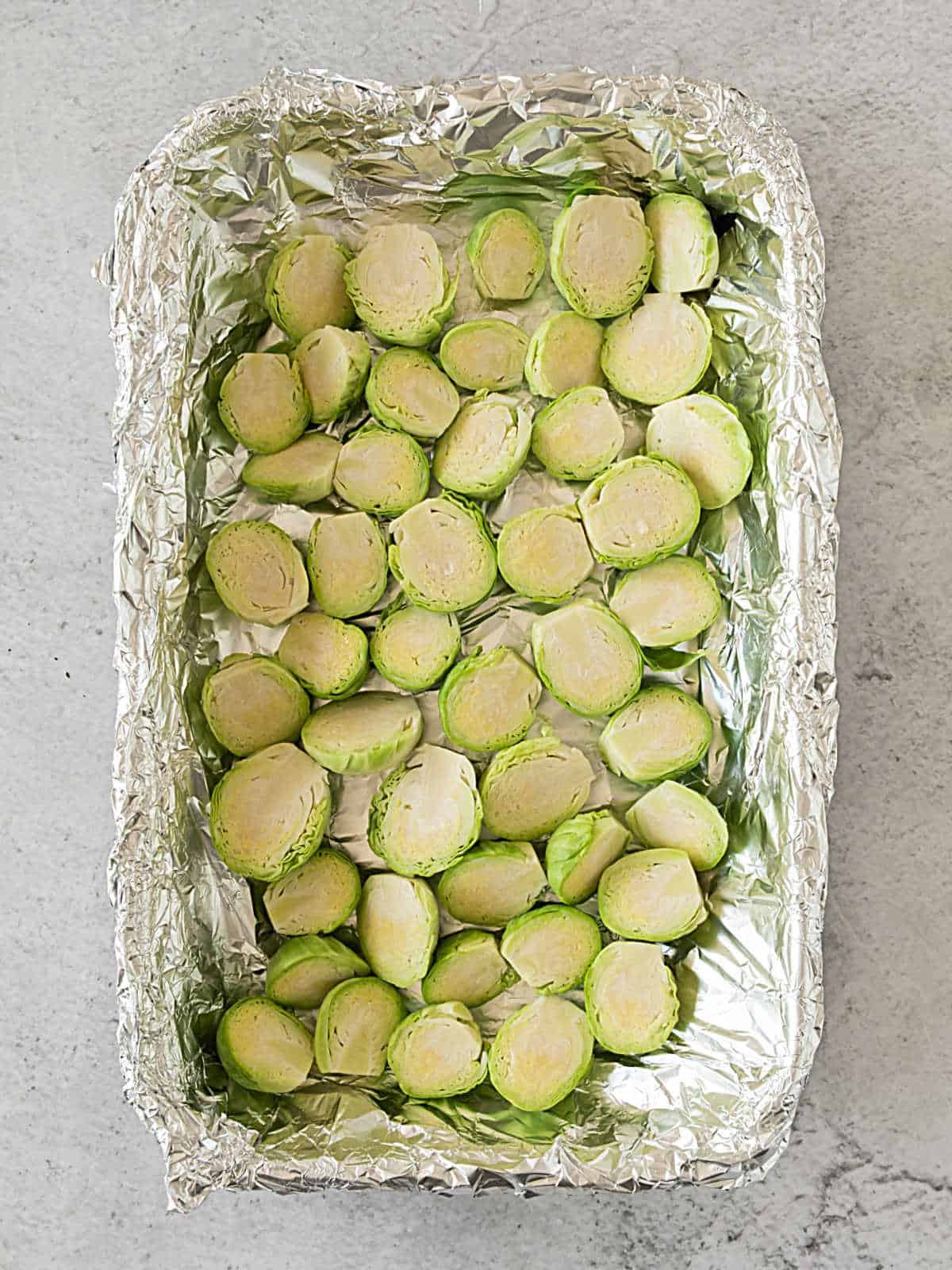 Halved raw Brussels sprouts in a foil lined pan on a light grey surface.