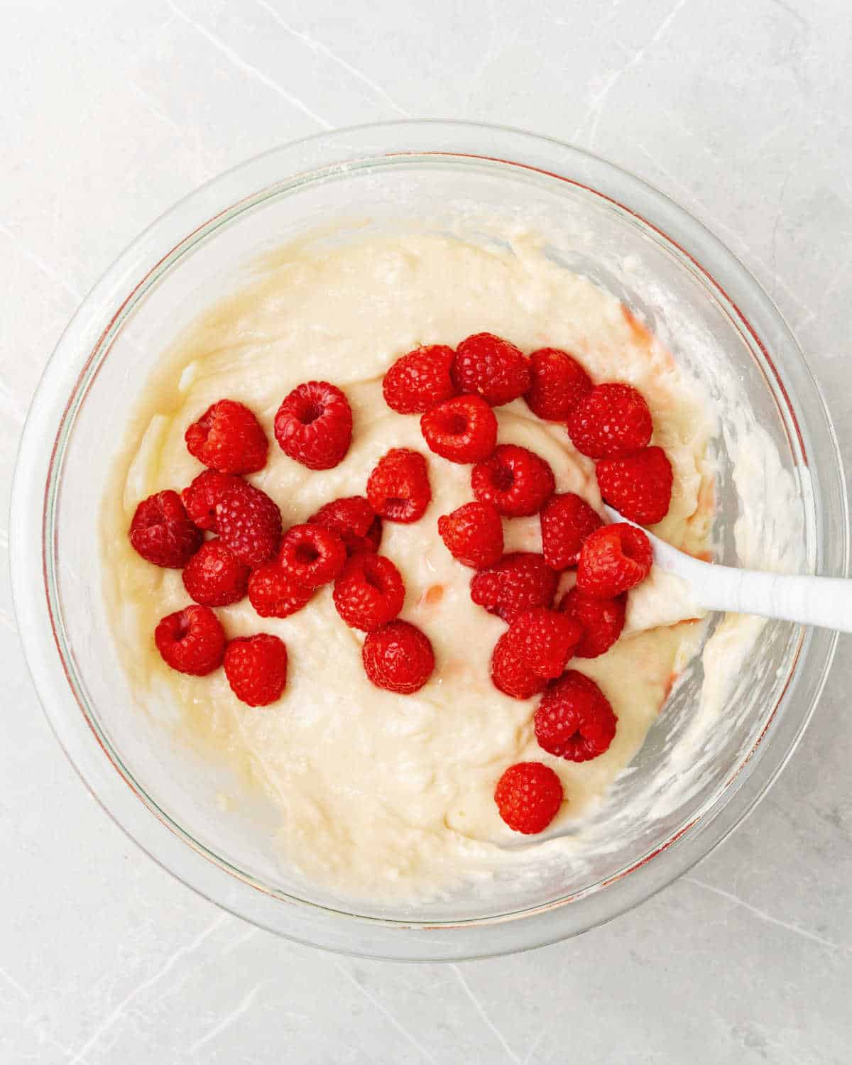 Fresh raspberries added to muffin batter in a glass bowl with a spatula. Light grey surface.