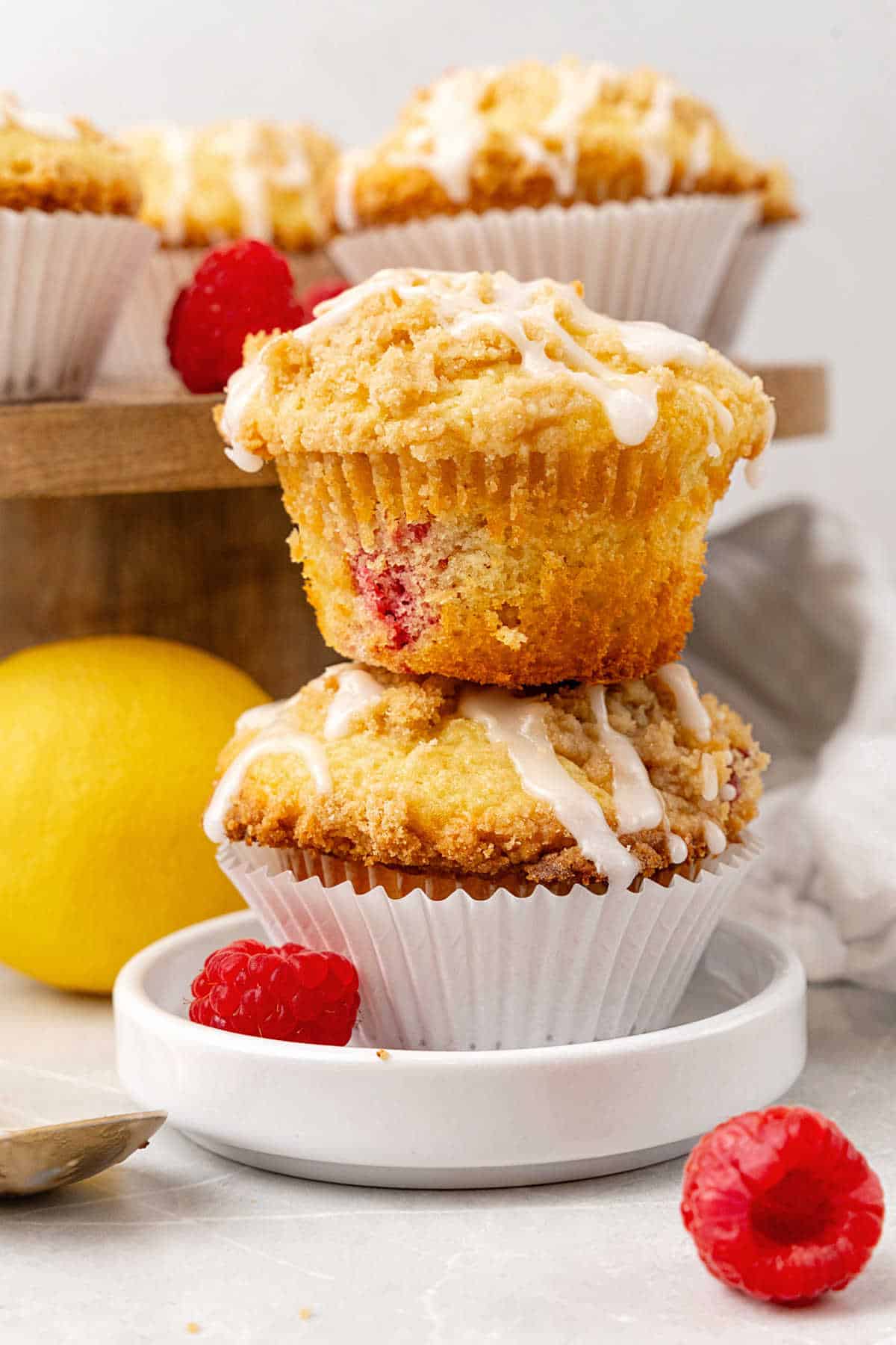 Two raspberry crumb muffins with glaze on a white plate. More muffins in background.