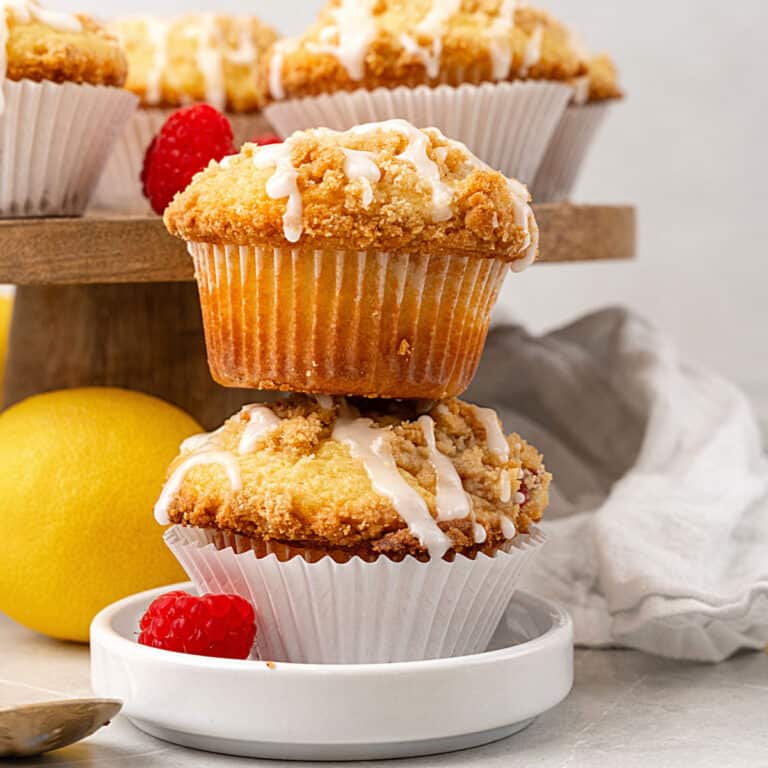 Stack of two glazed raspberry muffins on a white plate. Whole lemon and more muffins in the background.