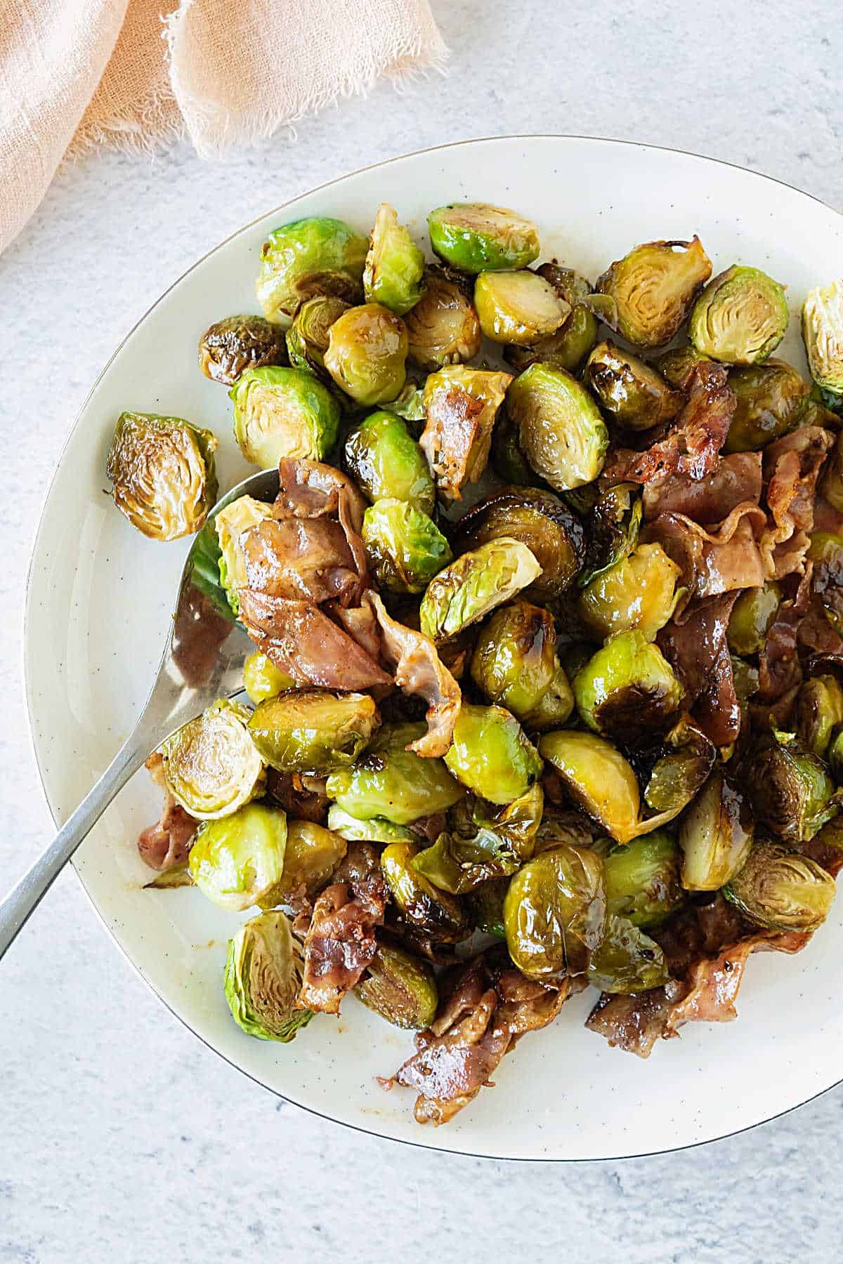 Top view of white plate with bacon roasted brussels sprouts. White surface, silver spoon. 