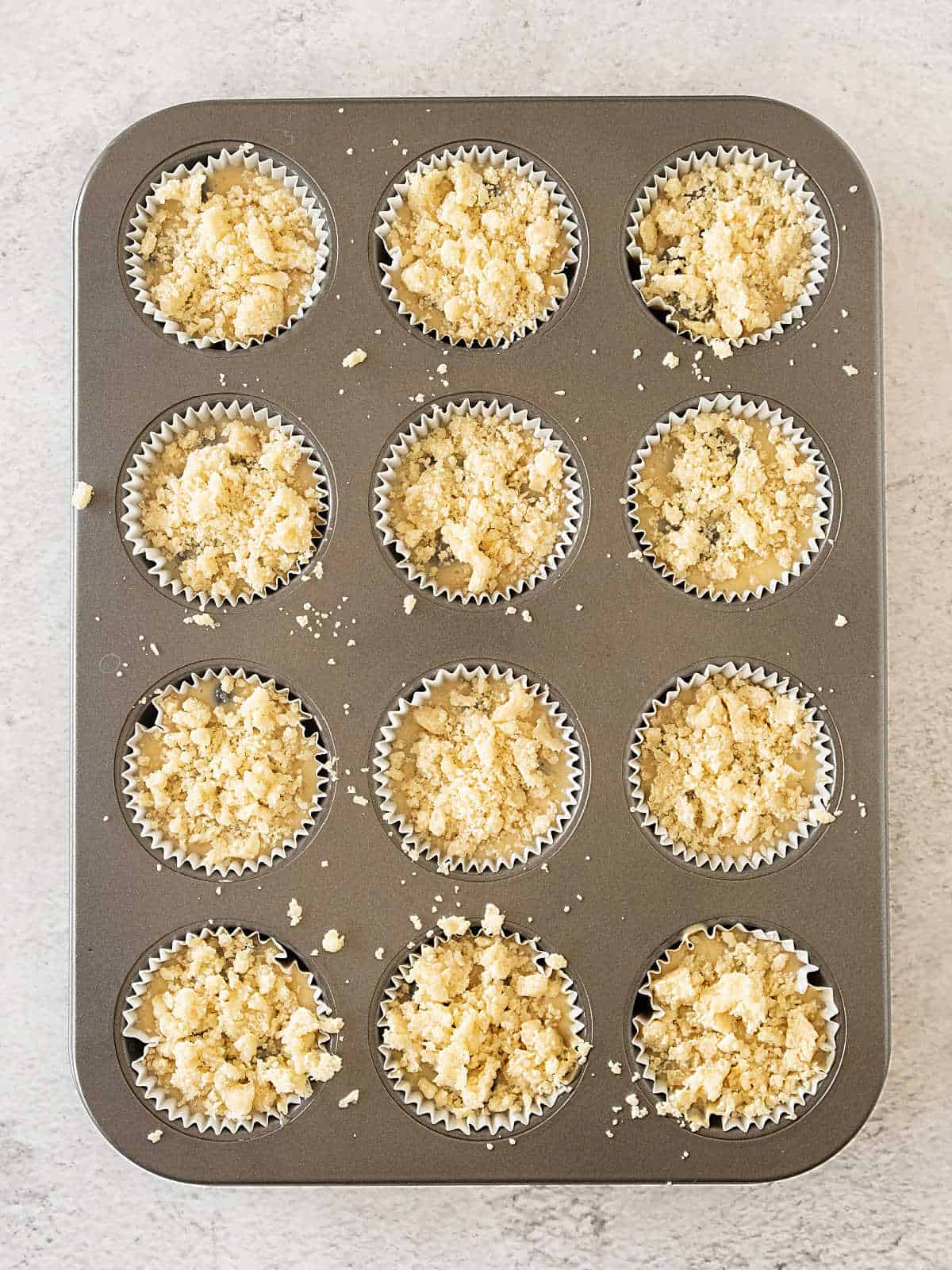 Crumble-topped muffins on the pan before baking. Light grey surface.