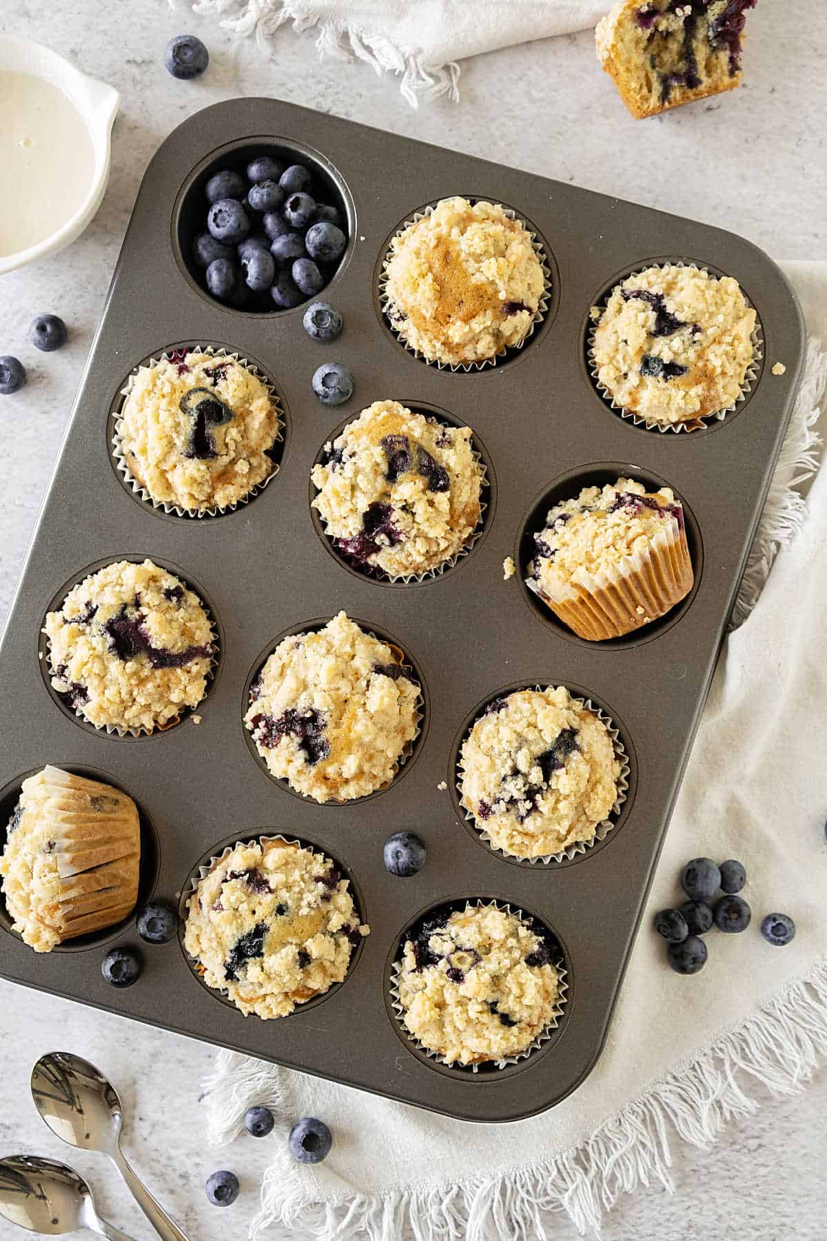 Metal pan with blueberry muffins with streusel topping. White surface with a white cloth.