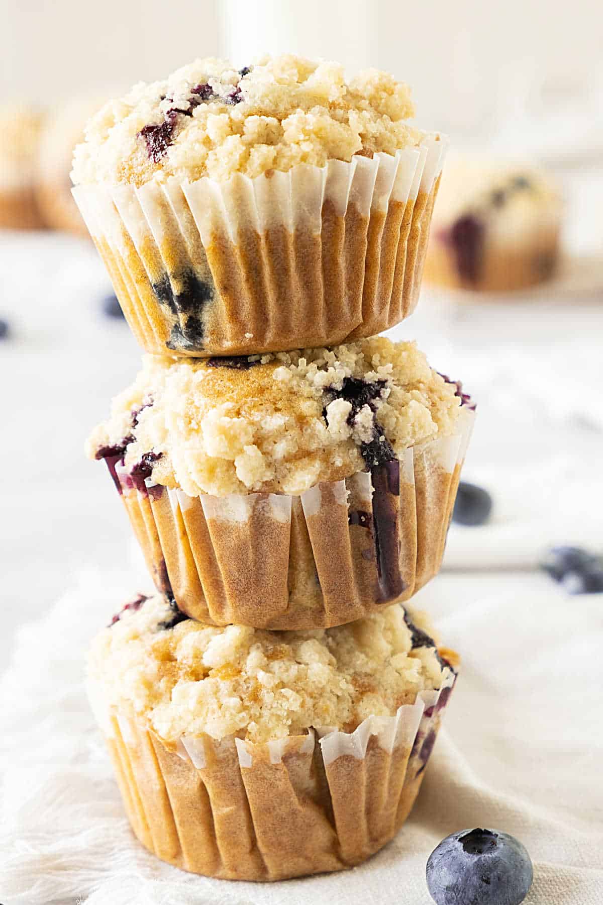 Stack of three blueberry crumb muffins in paper liners. White background. 