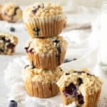 Three blueberry crumble muffins in a stack, one bitten beside them; white background with linens.