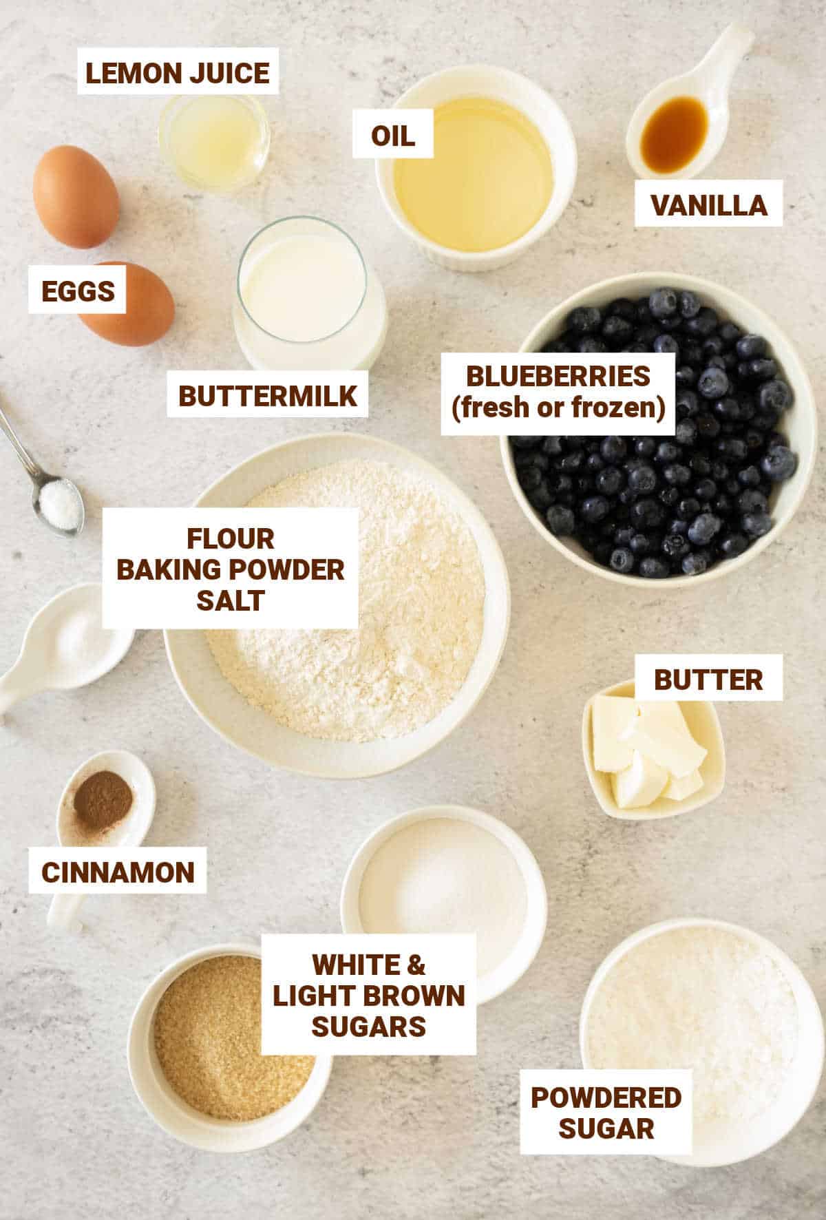 Ingredients for blueberry muffins in bowls on a grey surface including sugars, oil, butter, cinnamon, flour, eggs.