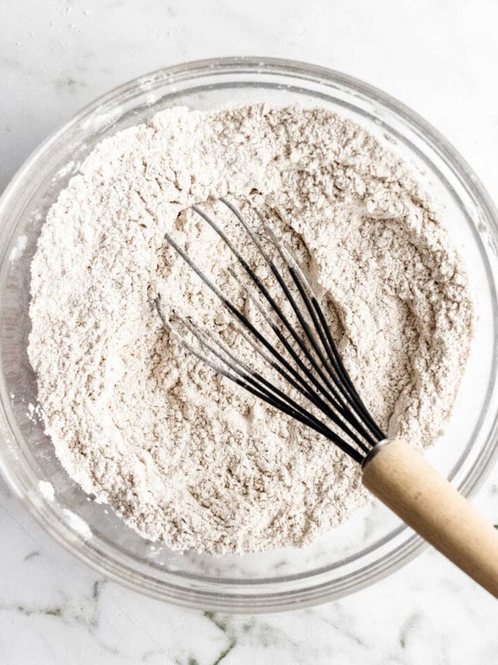 Flour in a glass bowl with a whisk on a white marble surface.