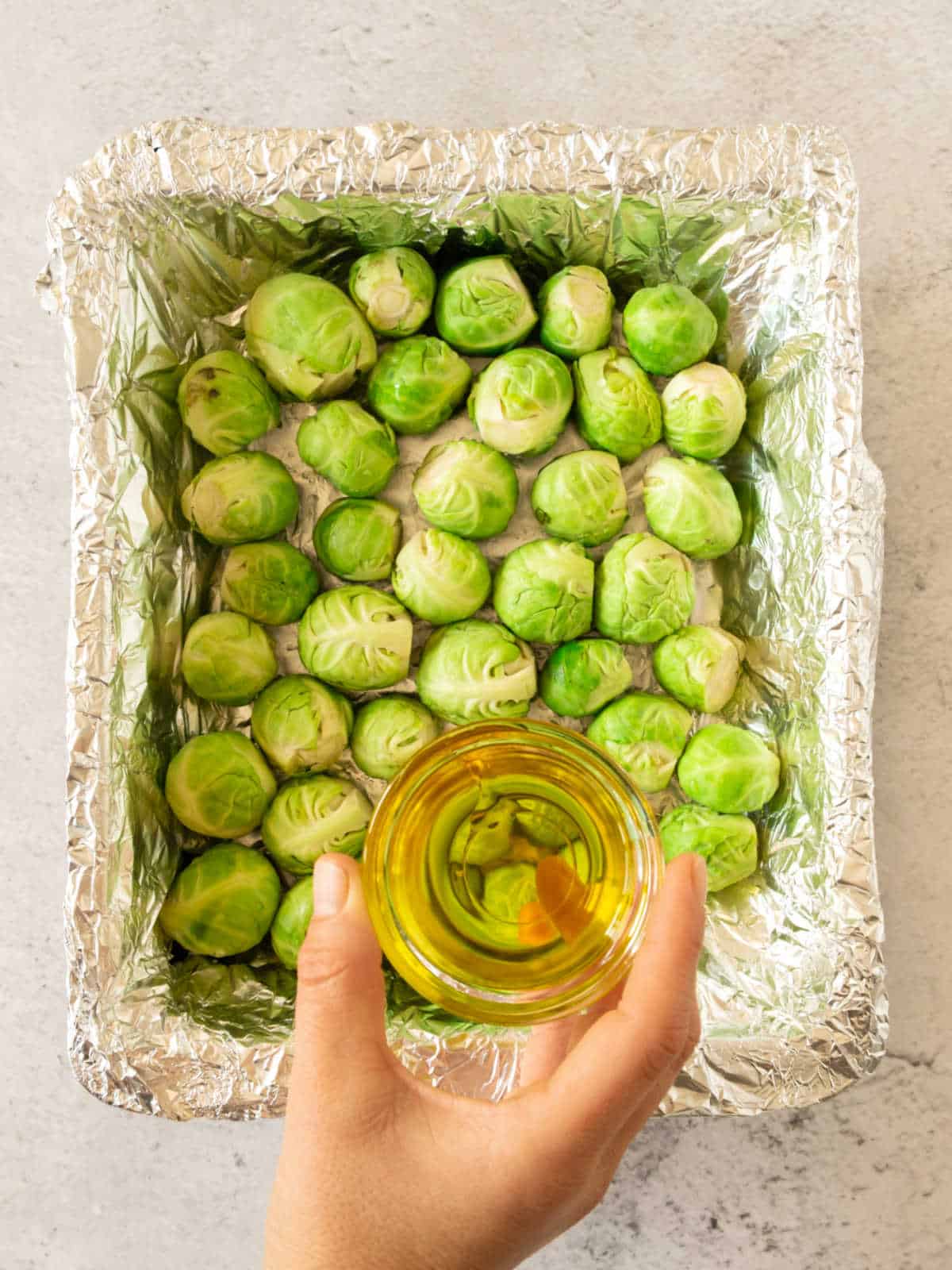 Hand with oil bowl above Brussels sprouts on a foil lined dish. Light grey surface.