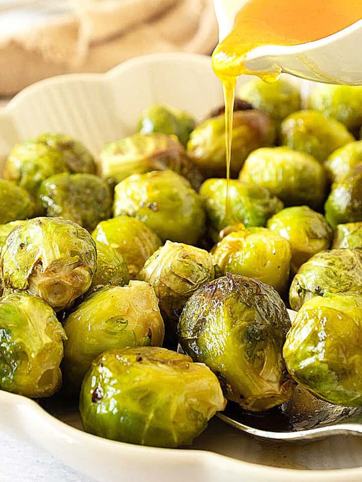 Drizzling honey glaze from a white jar over baked Brussels sprouts on a white platter.