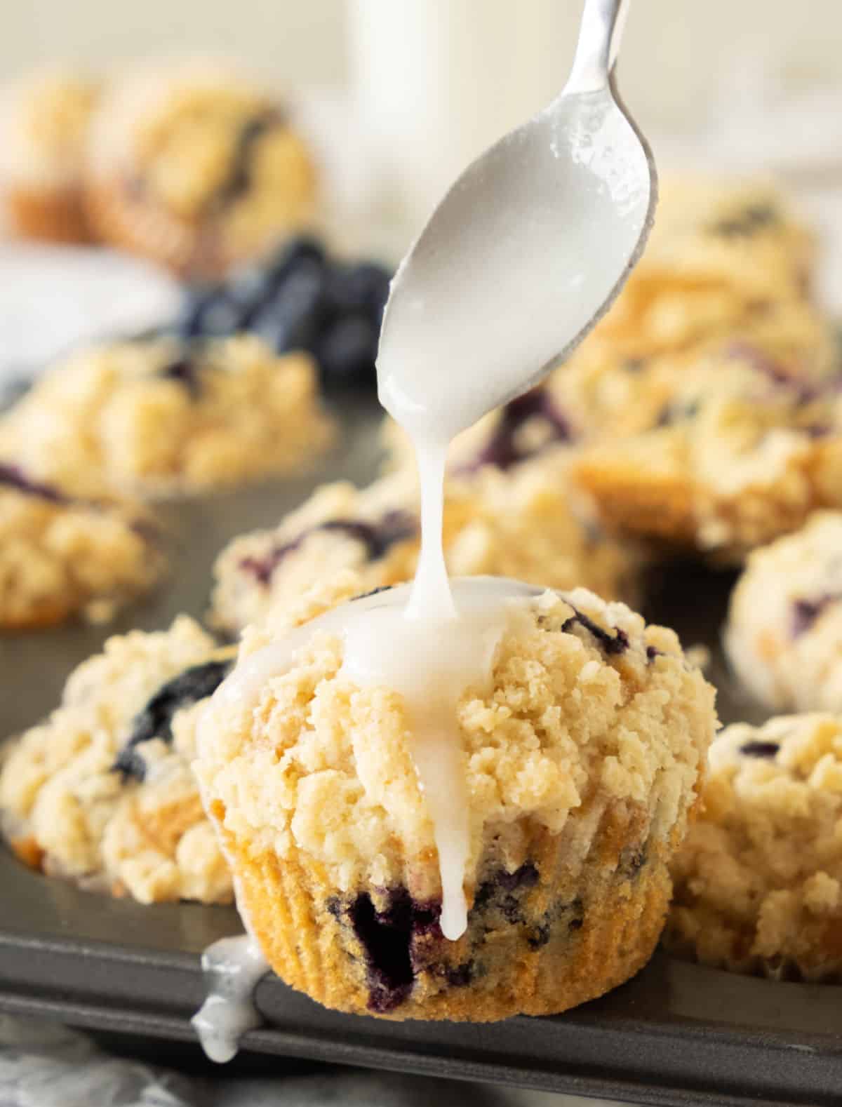 Glaze dripping from a spoon on a blueberry crumb muffin. More muffin in the background.