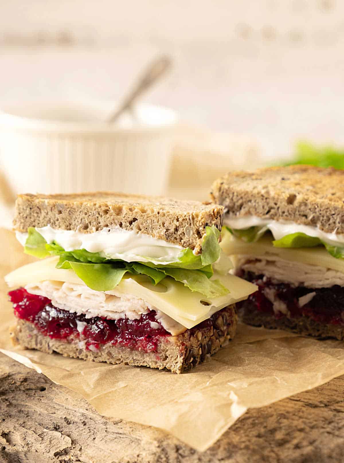Two halves of turkey, cranberry, lettuce sandwiches on a beige surface. White bowl in the background. 