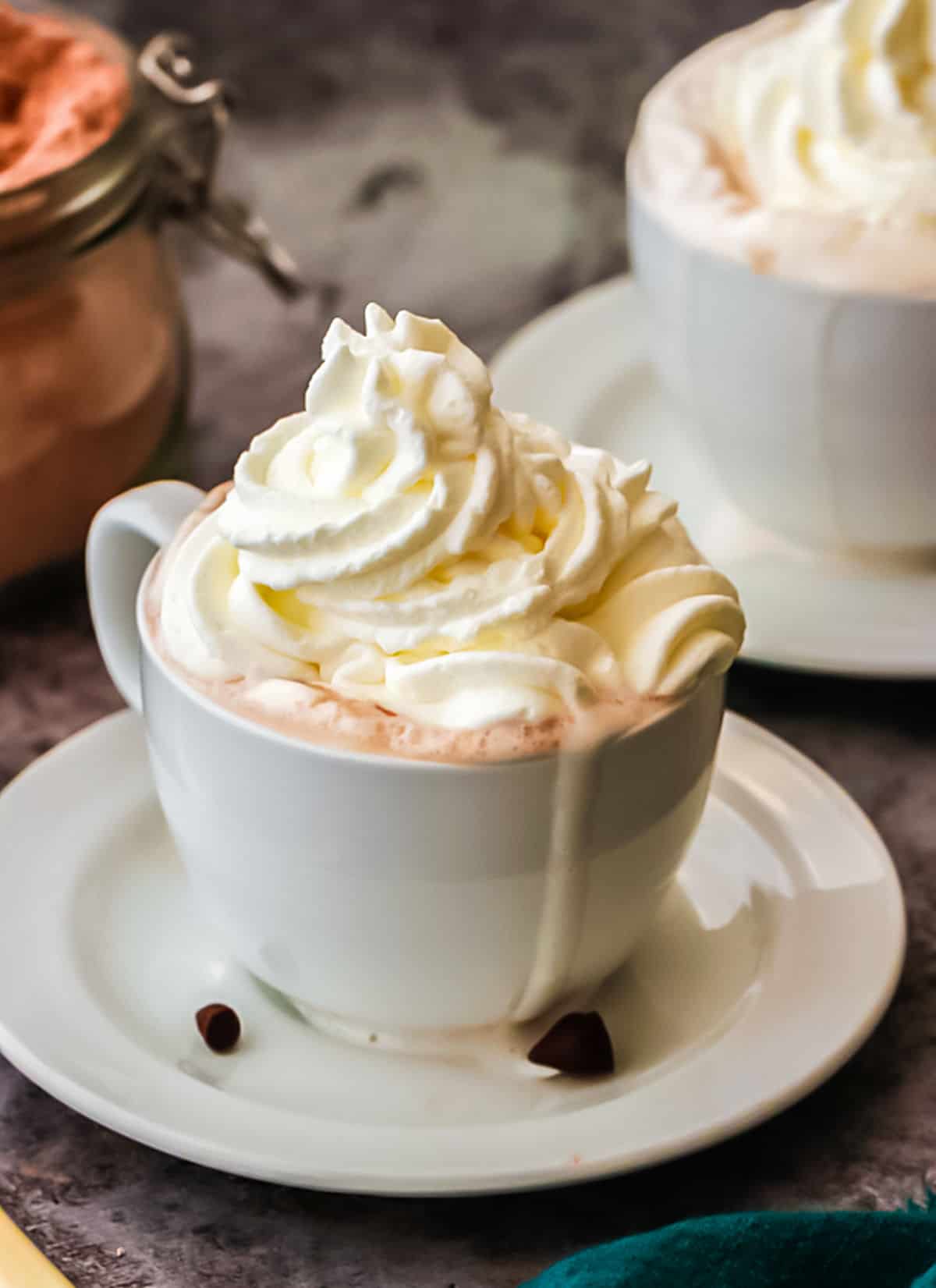 Whipped cream topped hot chocolate in a white cup with plate. Grey surface. 